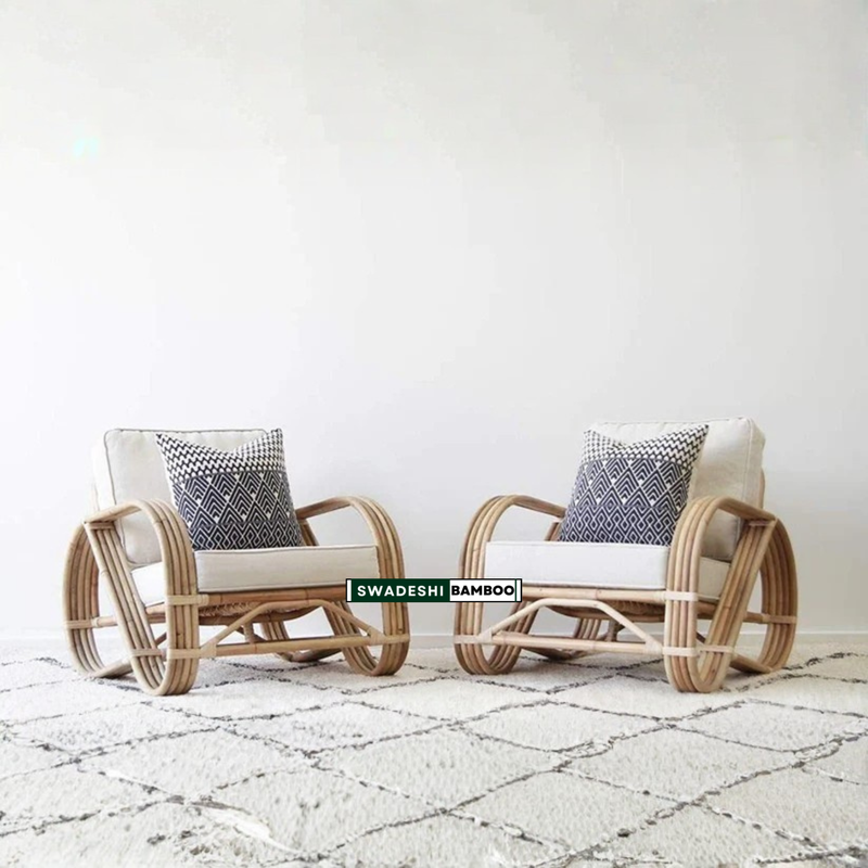 Two bamboo armchairs with cream cushions and patterned throw pillows, set on a textured white rug against a plain white wall. A green brand label reading 'SWADESH BAMBOO' is visible on the front of the chairs.