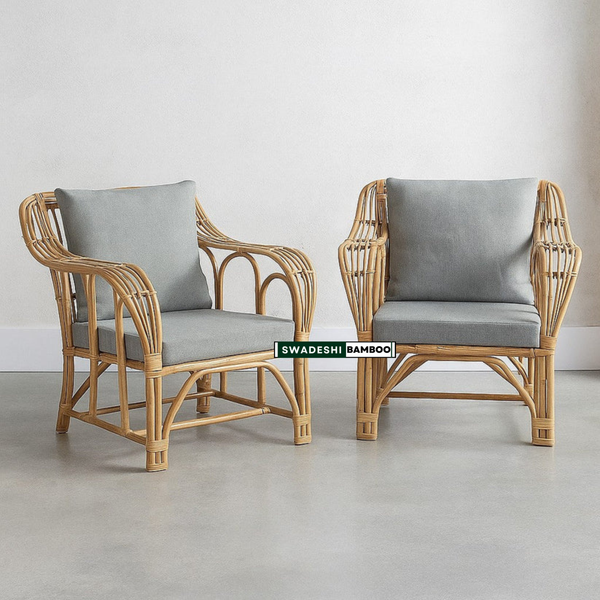 Two light natural bamboo armchairs with gray cushions, set against a minimalist gray wall and concrete floor. A green and white label in the center reads 'SWADESHI BAMBOO'.