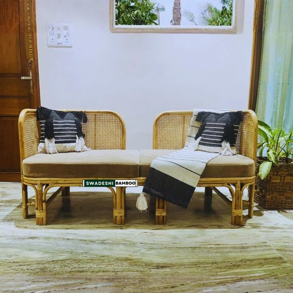 A two-seater bamboo rattan daybed with beige cushions, styled with two black and white striped pillows and a matching beige and black throw blanket. The daybed is placed on a textured beige and brown patterned rug in a home setting with a white wall, wooden door, and potted plant.